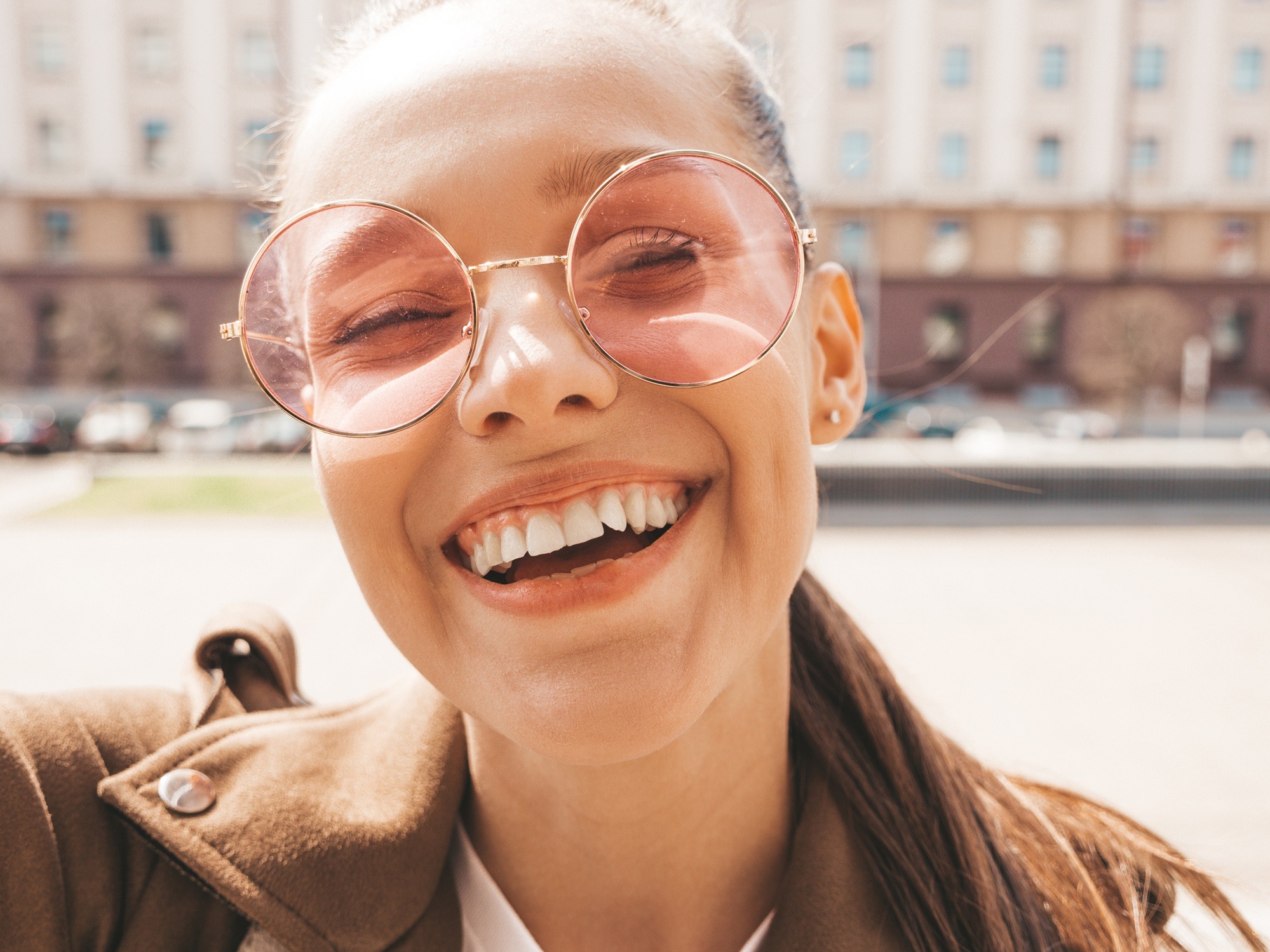 Closeup portrait of beautiful smiling brunette girl in summer hipster jacket. Model taking selfie on smartphone.Woman making photos in warm sunny day in the street in sunglasses