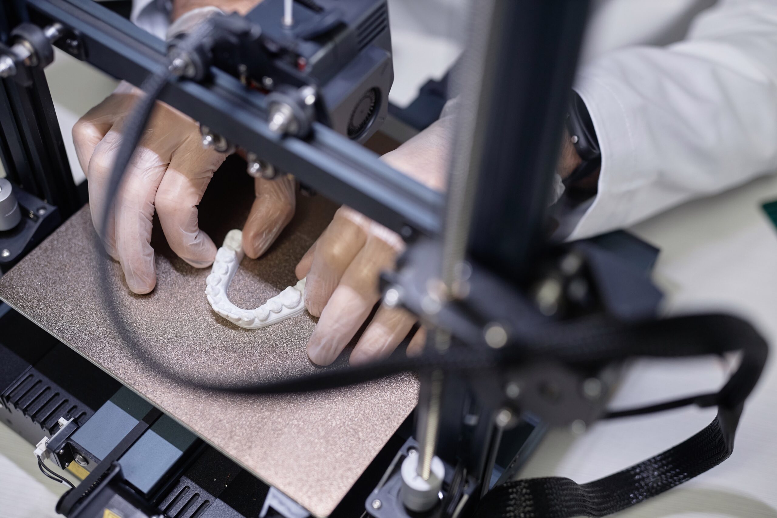 Top down view of illuminated while-colored dental model on aluminum 3D printer platform with unrecognizable technicians hands in gloves